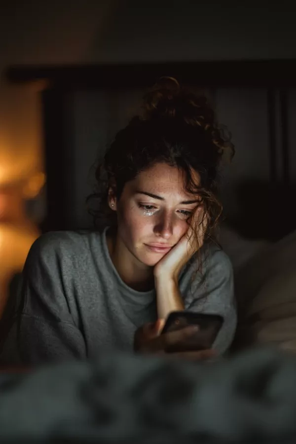 Sad woman in bed crying while looking at her phone in a dimly lit room